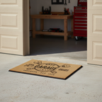 Dad's 24/7 Garage personalized coir doormat at an open garage entry with workbench and red tool chest
