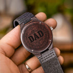 Hand holding an engraved wood watch showing DAD EST 2026 wreath design on the case back